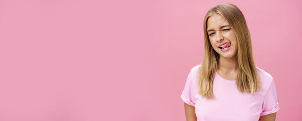 Playful flirty attractive young girl with fair straight hair in t-shirt winking at camera smiling and licking upper lip with tongue feeling enthusiastic and carefree against pink background - obrazy, fototapety, plakaty