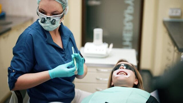 Woman lying on chair on cabinet tooth clinic with mouth dilator and protective goggles. Dentist in glasses with magnifying glass holding medical equipment. UV lamp for whitening or caries treatment.
