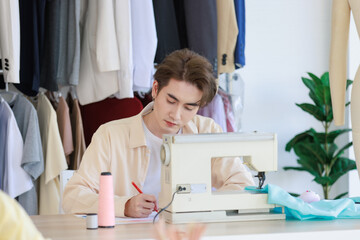 Millennial Asian young talent professional male dressmaker designer seamstress sitting working designing drawing sketching dress outline and choosing color pattern from collection at working desk
