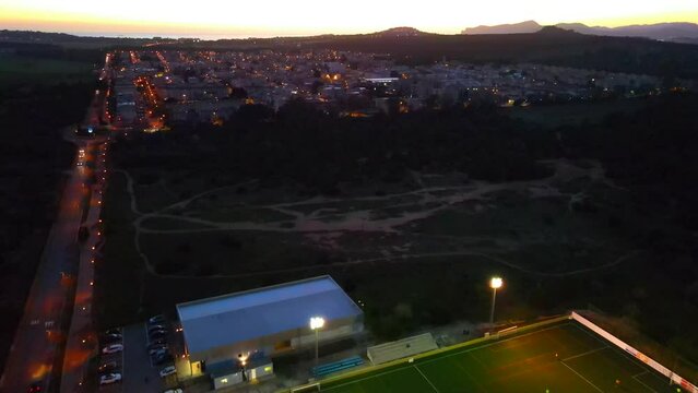 Backwards revealing drone shot of a lit up football sports field at dusk with a town in the background in Santa Ponsa, Mallorca, Spain.