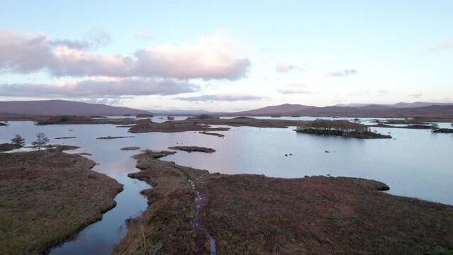 A drone slowly rises aup and tilts down towards a patchwork landscape of islands amongst fresh water lochs (lakes) and peat bogs at sunset. Loch Ba, near Glencoe, Scotland.