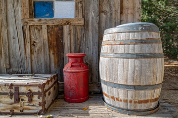A barrel, a metal jug, and a trunk on the porch of an old wood cabin