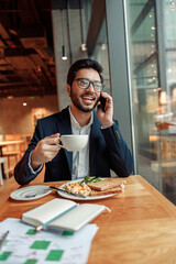 Happy indian businessman is talking phone with client during lunch time in cozy cafe