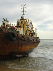 Fototapeta premium Old rusty tugboat on beach was run aground, stranded and abandoned, with bow in ocean waves in portrait view.