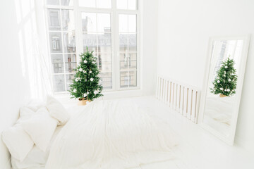 Comfort, home and Christmas Eve concept. Bedroom with white soft bed, mirror, big window for light coming in room, decorated New Year tree and ladder. Holiday decoration.
