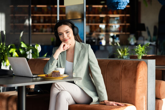 Buisness woman in modern new headphones wearing elegant suit sitting at caffe in front of computer laptop having break after hard day work.