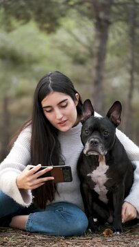 Young caucasian woman taking a selfie hugging her american staffordshire and french bulldog mixed breed dog at a pine forest.