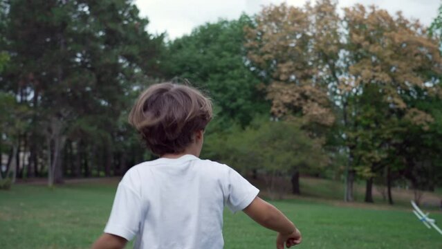 Back of happy child throwing foam glider plane. One small boy runs outdoors with airplane toy at park