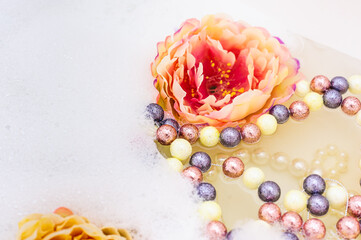 Multicolored beads and pearl necklace next to peony flowers float in water with foam. copy space. close up