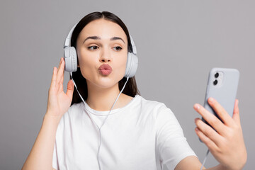 Adorable cute young woman in basic white t-shirt lsending kisses at cellphone smartphone wearing headphones and earphones