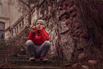 Sad guy in autumn sits on steps against background of building ruins, around dried shoots of plants, fallen foliage. Boy looks thoughtfully into the sky, his head propped up with hand, view from below