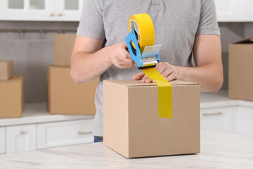 Man taping box with adhesive tape dispenser in kitchen, closeup