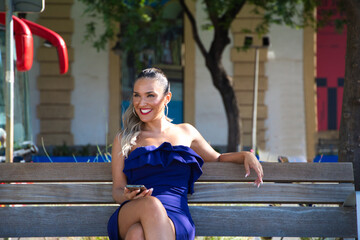 Young and beautiful blonde latina woman dressed in short dress is resting sitting on a bench in the city. The woman is consulting her mobile phone.