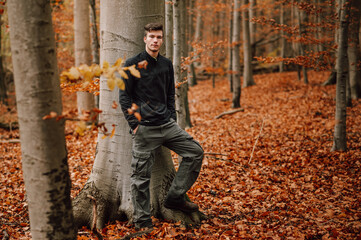Junger Mann im Wald im Herbst, in ländlicher Natur mit Herbstlaub in Buchenwald. Lehnt an einen Baum und sieht in die Kamera, zum Betrachter. Im Vordergrund unscharfes Herbstlaub