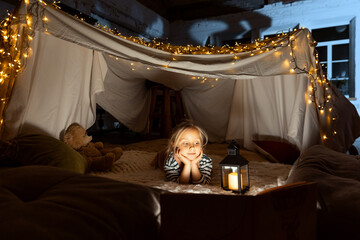 Fototapeta na wymiar Cute little girl, child, daughter lying inside hut, tent with lights, toys and pillows and reading book. Playful, cozy evening