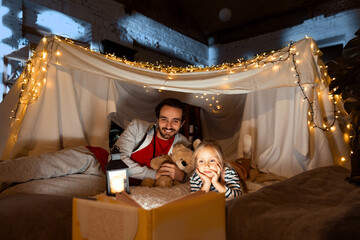 Fototapeta na wymiar Cheerful father and daughter lying inside self-made hut, tent in room in the evening and reading book. Fairy tales before sleep
