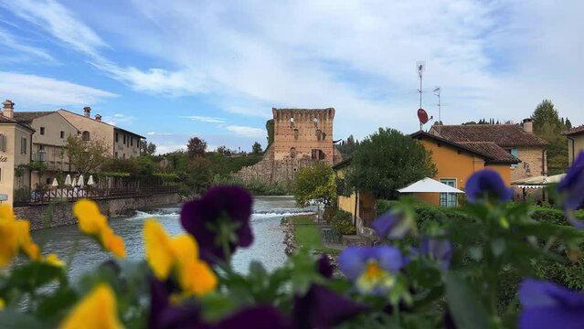 Scenic view through yellow and blue colorful flowers of Borghetto sul Mincio charming ancient Italian hamlet in Veneto, Italy