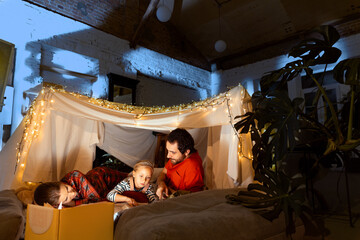 Fototapeta na wymiar Happy family, mother, father and daughter lying inside self-made hut, tent in room in the evening. Cozy time with parents and child