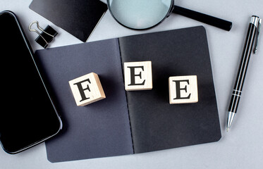 Word FEE on wooden block on black notebook with smartpone, credit card and magnifier