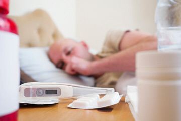 Pills and electric thermometer in focus. Bald sick man out of focus in the background. Flu and winter season infection concept.