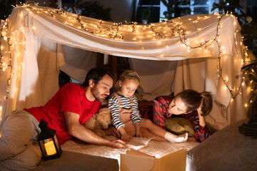 Obraz na płótnie Canvas Happy family, mother, father and daughter lying inside self-made hut, tent in room in the evening and reading book. Merry Christmas