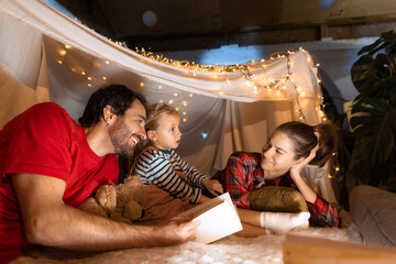 Mother, father and daughter lying inside self-made hut, tent in room in the evening and reading book. Having fun