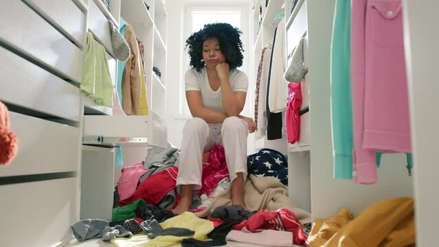 Cute African American woman with curly hair frustrated in closet 4K. Slow motion of funny sad woman of color sitting in the middle of wardrobe looking around at clothes mess, doesn't know what to wear