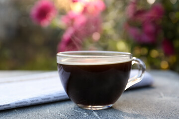 Glass cup with coffee and newspaper on grey table outdoors, closeup. Morning ritual