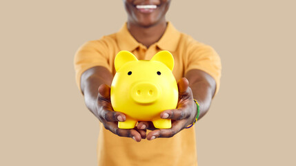 Piggy bank in form of yellow pig in hands of smiling black man on beige background. Close up of...