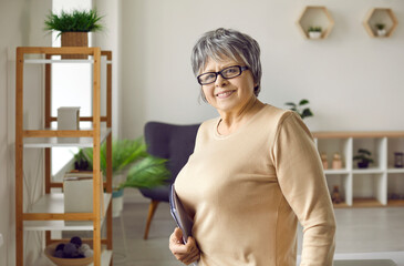 Portrait of smiling cute senior woman with notepad in hand in living room of her home. Happy gray-haired mature woman who likes to write to-do list in notebook posing smiling at camera.