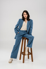 full length of young woman in blue and stylish suit holding hand in pocket while posing on stool on...