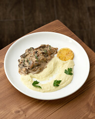 Fried mushrooms in creamy sauce with mashed potatoes on white plate. Dish on wooden table. Dark blurred background. Top view, close-up.
