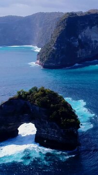 Vertical aerial video of island with half donut shape and waves crashing against it