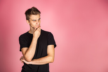 Pensive doubtful man thoughtful at pink background, touching chin, thinking, looking away. Serious young guy in casual t-shirt deep thinking. Advertising information concept. Copy text space