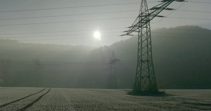 W&uuml;rgassen, Germany - 12/17/2022: High-voltage pylons with overhead lines against the sun in the evening in the wind on a field with ice