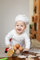 An Asian baby in a chef's cap and apron bakes Christmas ginger cookies he smiles happily and looks at the camera
