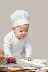 An Asian baby in a chef's cap and apron, smeared in flour, prepares Christmas ginger cookies and she cries because something doesn't work out