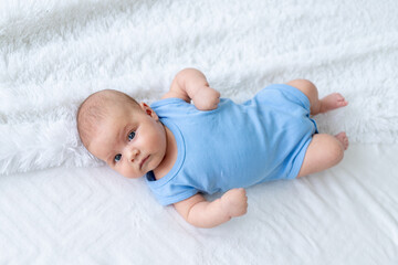 cute baby boy three months old in a blue bodysuit on a white bed at home