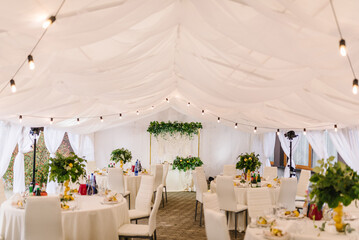 Wedding setup. The wedding arch is decorated leaves and a garland with lights in tent. Luxury elegant tables setting dinner and chairs.  Banquet hall with decor flowers, herbs, citrus fruits lemons.