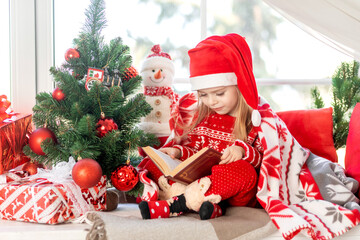 a cute girl child is reading a book sitting on the windowsill at the window of the house at the Christmas tree and waiting for the new year or Christmas in a red Santa Claus hat and smiling