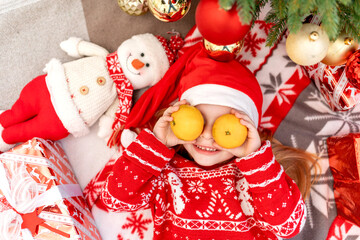 a happy baby girl lies under the Christmas tree at home with tangerines and waits for the new year or Christmas in a red Santa Claus hat and smiles and rejoices