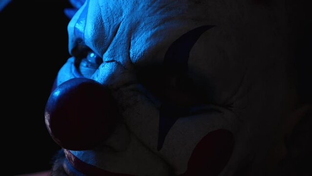 Close-up of the face of a scary evil clown in a dark room with blue lights. A man wearing a scary clown mask in the dark. His gaze is directed at the camera