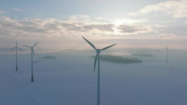 Aerial establishing view of wind turbines generating renewable energy in the wind farm, snow filled countryside landscape with fog, sunny winter evening golden hour, wide drone shot moving forward