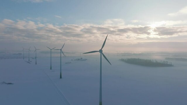 Aerial establishing view of wind turbines generating renewable energy in the wind farm, snow filled countryside landscape with fog, sunny winter evening, golden hour light, wide drone orbiting shot
