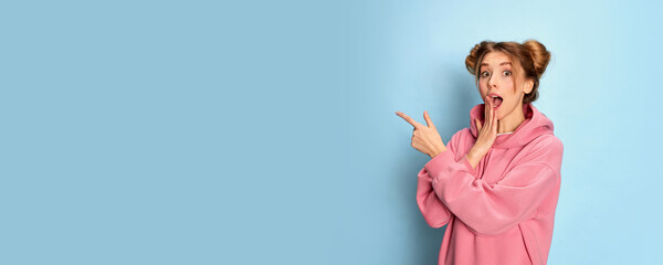 Portrait of young emotive girl in pink hoodie posing, pointing with shocked face over blue studio...