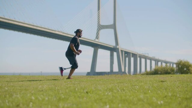 Tracking shot of athlete with prosthetic leg jogging in park on summer day. Man with disability running along river bank with bridge in background. Amputee sport, motivation concept.