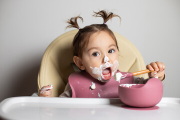 Small cute little toddler brunette caucasian girl with two tails eating by herself with a spoon the...