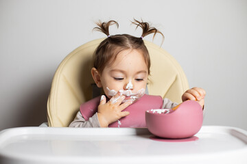 Small cute little toddler brunette caucasian girl with two tails tasting by herself with a spoon the greek yogurt sitting in baby chair with messy face; self-feeding concept