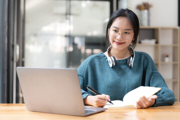 Young adult happy smiling Asian student wearing headphones talking on online chat meeting using laptop in university campus or at virtual office. College female student learning remotely.