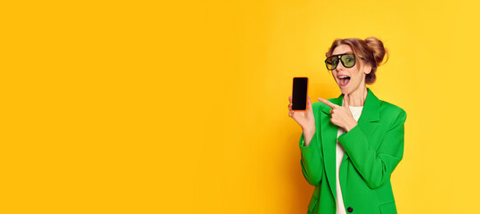 Portrait of young girl in bright green jacket posing, pointing on phone screen with excitement over yellow studio background. Online shopping